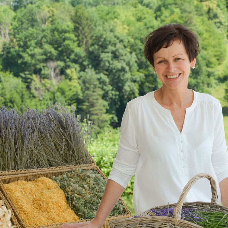 Eine Frau Präsentiert frische Kreuter in Körben, im Hintergrund ein Waldstück mit Wiese